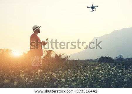 Silhouette of a man photographer is using RC aerial drone for video shooting, while is standing in rural against beautiful sunset. Male tourist is taking photo on flying quad-copter during summer trip