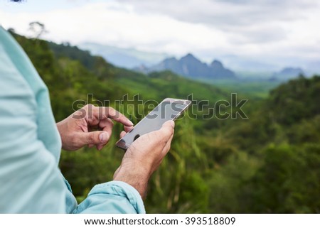 Closeup of a man\'s hand is holding mobile phone with copy space screen against blurred jungle landscape. Young male is searching information on cell telephone during summer adventure in Thailand