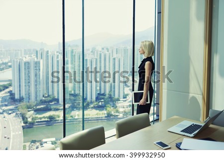 Blonde woman proud CEO is holding touch pad and looking in office window with cityscape, while is standing in conference room near table with open laptop computer during her business trip in New York