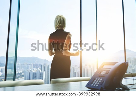 Back view of a businesswoman with digital tablet in hands is watching in office window on a New York city view and thinking about future her company. Female manager is using touch pad during work day