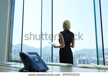 Woman proud CEO with digital tablet in hands thoughtful is looking into the office window, while is standing in skyscraper interior in big conference room with table. Copy space for advertising text