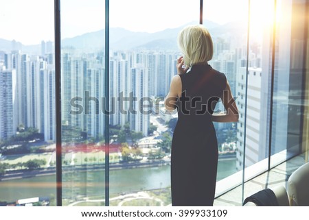 Back view of young entrepreneur is calling via cell telephone, while is standing in her private office against window with view of developed business district with high skyscrapers.Copy space for text