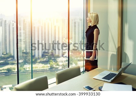 Woman skilled employer with touch pad in hand is looking in office skyscraper window of conference room while is thinking that adopt a decision after interview with specialists in the economic sphere