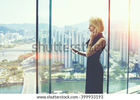 Young proud businesswoman is talking on her mobile phone with client and reading order on digital tablet, while is standing near office window with view of developed business district in China