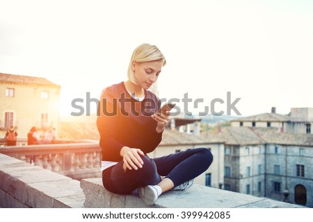 Female tourist is searching information on website via mobile phone about history this beautiful old town. Hipster girl is reading text message on cell telephone, while is sitting on a building roof