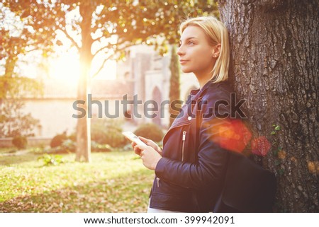 Young female student with cell telephone in hands is dreaming about something, while is standing on a campus. Thoughtful woman tourist is holding smart phone, while is enjoying free time in vacation