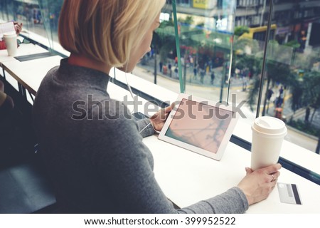 Young woman is watching movie in network on touch pad,while is sitting with take away coffee in cafe with city view outside window. Hipster girl is holding digital tablet with copy space on the screen