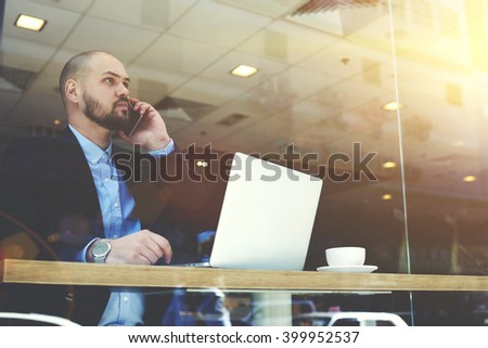 Young bearded man CEO of big successful company is having mobile phone conversation with customers, while is sitting with portable laptop computer in modern restaurant interior during work break