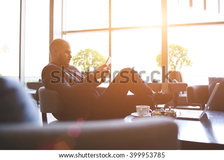 Young man confident entrepreneur is reading financial news via internet on portable touch pad, while is relaxing in cozy cafe. Male proud CEO is using digital tablet, while is waiting partner in bar