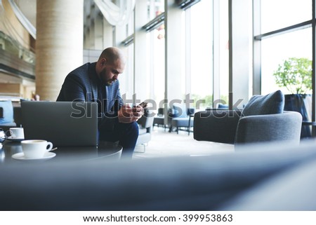 Man professional banker is reading text message on mobile phone, while is resting after work on laptop computer. Male entrepreneur is using cellphone, while is waiting business lunch in restaurant