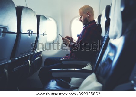Man is listening to music in headphones and chatting in social network via mobile phone, during his flying in an airplane. Hipster guy is watching video on cell telephone , while is sitting in plane
