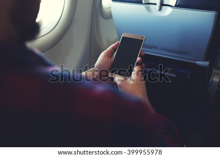 Close up image of man`s hands is holding cell telephone with blank copy space screen for your promotional content. Hipster guy is reading text message on mobile phone during flying in an airplane