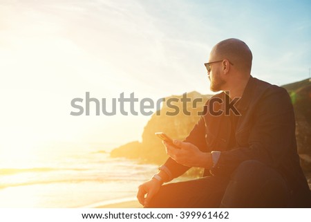 Young hipster man traveler with cell telephone in hand is enjoying amazing landscape, while is sitting on the beach near ocean with copy space background for your text message or advertising content