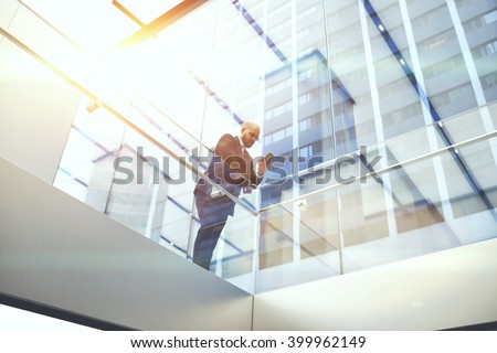 View from below of businessman is reading financial news on web page via cell telephone, while is standing in modern skyscraper interior. Confident male CEO is using mobile phone during work break