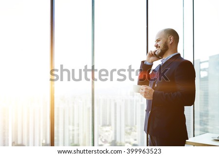 Smiling happy man managing director talking on mobile phone about his successful career development, while is standing with cup of coffee in hand in his office near window background with copy space