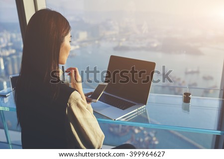 Woman owner of successful company is using mobile phone during video conversation in internet via net-book with staff during her business trip in New York, while is sitting in skyscraper building