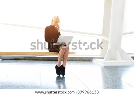 Woman designer of websites is talking on mobile phone with customer, while is sitting with laptop computer in co-working space. Pretty female with net-book on knees having cell telephone conversation