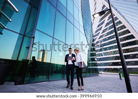 Young successful businessman in luxury suit is reading monthly report on portable digital tablet, while his female secretary with mobile phone and folder documents in hand is commenting her work