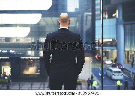 Back view of man proud CEO in luxury suit is watching at big office window at central street of New York, while is resting after important meeting with international partners during business trip