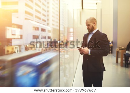Bearded man skilled managing director is searching on web site via mobile phone needed information for meeting with clients, while is standing near window with view of the main street of New York