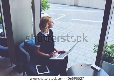 Woman is looking on street outside the window, while is sitting with net-book and touch pad in luxury restaurant. Young female is thinking about something after learning on-line via laptop computer