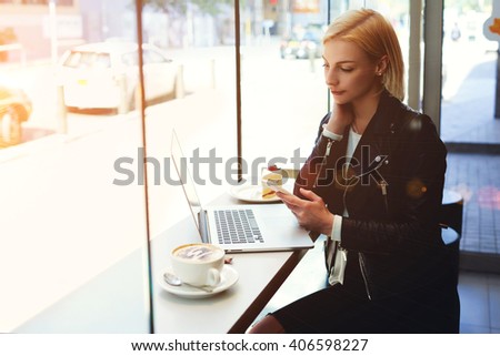 Hipster girl is reading text message on cell phone, while is watching on-line training in network via laptop computer. Business woman is sitting in coffee shop with cup of cappuccino during work break