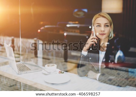 Woman freelancer is talking on mobile phone with her boyfriend, while is sitting in urban coffee shop at the table with open portable laptop computer, cup of cappuccino and dessert. Female student