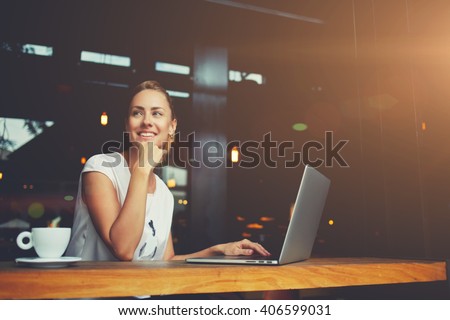 Young female with cute smile sitting with portable net-book in modern coffee shop interior during recreation time, charming happy woman student using laptop computer to prepare for the course work
