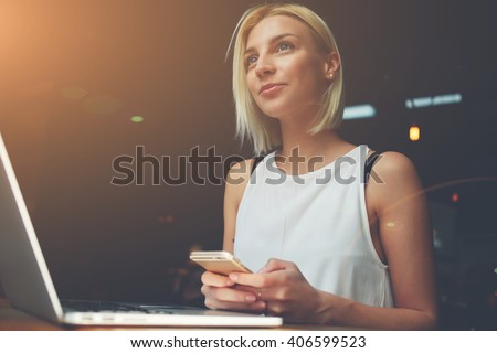 Young beautiful blonde female holding mobile phone while sitting with portable net-book in coffee shop interior, charming dreamy woman using cell telephone and laptop computer during rest in cafe
