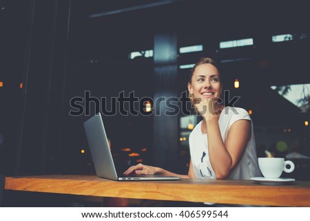 Young female with cute smile sitting with portable net-book in modern coffee shop interior during recreation time, charming happy woman student using laptop computer to prepare for the course work