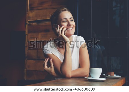 Young happy woman talking on mobile phone with friend while sitting alone in modern coffee shop interior, smiling hipster girl calling with cell telephone while relaxing after walking in summer day
