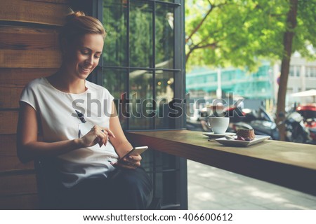 Charming woman with beautiful smile reading good news on mobile phone during rest in coffee shop, happy Caucasian female watching her photo on cell telephone while relaxing in cafe during free time