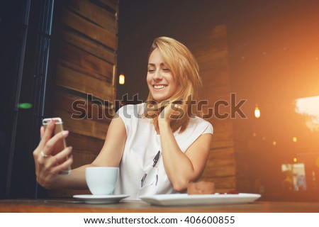 Young cheerful woman posing while photographing herself on smart phone camera for a chat with her friends, attractive smiling hipster girl making self portrait on cell telephone while sitting in cafe