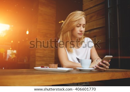 Young Caucasian female reading text message on her mobile phone while sitting in modern coffee shop interior, gorgeous hipster girl watching video on cell telephone during morning breakfast in cafe