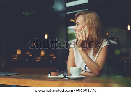 Young happy hipster girl speaking with her friend by mobile phone while relaxing in cozy coffee shop, charming lady with beautiful smile talking on cell telephone while sitting in cafe during lunch