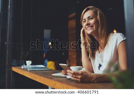 Happy female using smart phone while relaxing in cafe after walking during her summer weekend, charming smiling hipster girl received good news on cell telephone while she sitting in cozy coffee shop