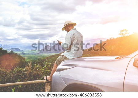 Young man is looking way on navigation via cell telephone during his amazing summer adventure in Thailand. Bearded male is watching photos on smart phone, while is sitting on a suv hood against jungle