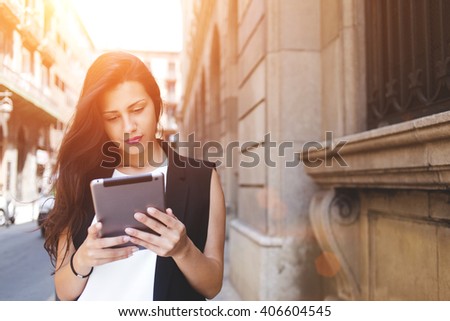 Beautiful woman is reading text message on touch pad while is standing on the street in spring day, stylish hipster girl tourist is using tablet computer for navigation during walk in the city abroad