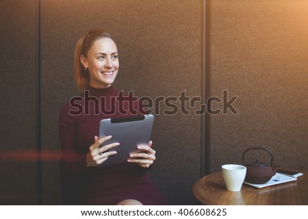 Beautiful happy woman enjoying good electronic book while is sitting at the table with cup of tea in modern coffee shop, smiling female thinking about something positive during work on digital tablet