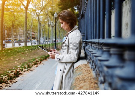 Young beautiful woman is booking on-line via portable touch pad, while is standing on the street in autumn day. Female dressed in spring clothes is reading information in internet via digital tablet
