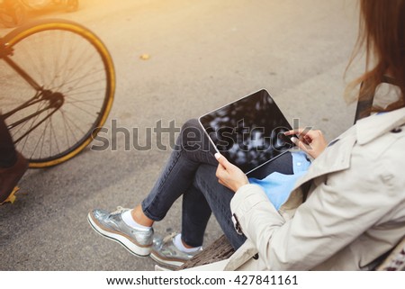 Rear view of young hipster girl is using portable touch pad for playing games, while is waiting car on a bus stop. Closeup image of stylish woman is holding digital tablet with copy space on screen