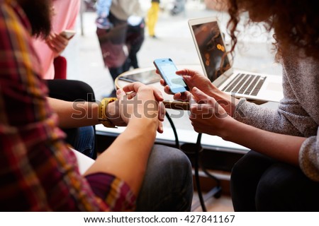 Closely image of woman is making an on-line purchase via cell telephone, while is sitting with friends in hipster cafe interior. Young female is reading information on web page via her mobile phone