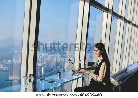 Woman start up owner of big prosperous enterprise is working on laptop computer during her business trip to China, while sitting in coffee shop in skyscraper near window with view of metropolitan city