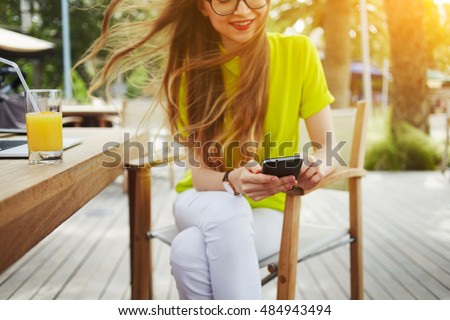 Smiling hipster girl is reading pleasant text message from her friend on mobile phone, while is sitting in cafe. Cheerful girl is watching funny video on cell telephone during free time in coffee shop