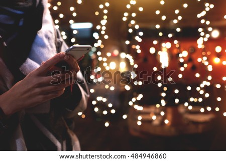 Closely image of young hipster girl is using her smart mobile phone, while is standing on the street against christmas garlands lights. Woman chatting with friends in social network via cell telephone