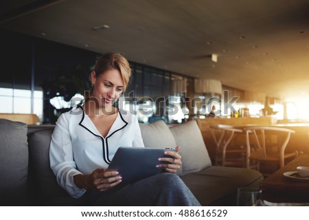 Female financier is reading financial news in internet via touch pad during work break in modern cafe. Confident woman lawyer is using digital table, while is waiting client in an informal setting