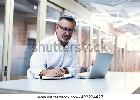 Experienced male skilled economist writing notes for delegate projects to his subordinates. University professor in eye glasses prepare for lectures after reading electronic book on laptop computer