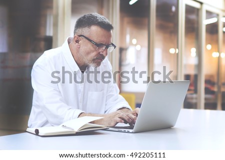 Skilled man programmer develops application software meanwhile it uses laptop computer and a notebook to record-keeping. Handsome mature businessman in eye glasses is sending e-mail by using net-book