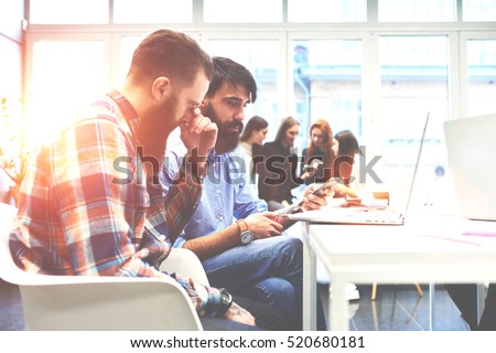 Bearded hipsters are involved into process of startup project creating in coworking space. Handsome professional is showing new app on smartphone.On the background four beautiful girls browsing photos