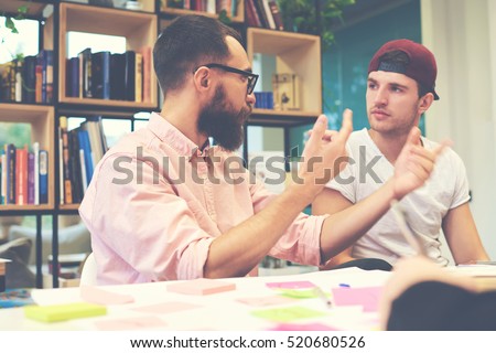 Two male hipsters architects sharing business thoughts ideas sitting in office.Young students preparing for making report by using special literature in library. Casual dressed teacher talk to pupil